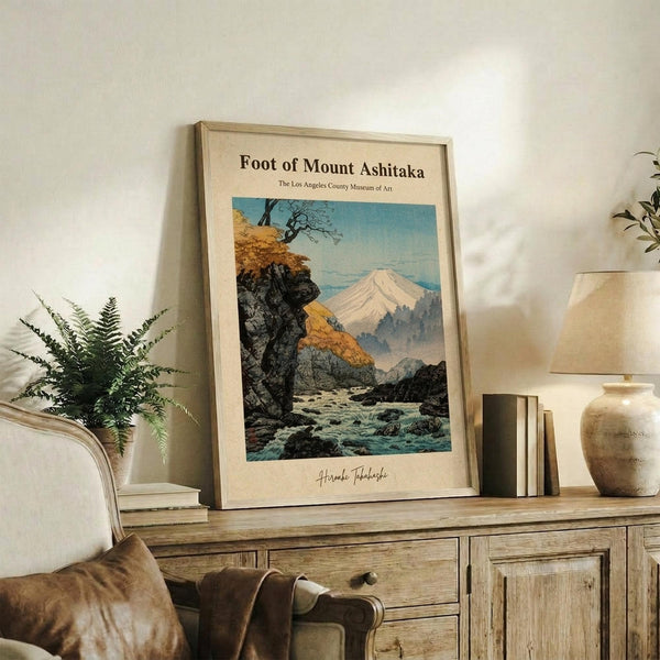 May include: Vintage Japanese woodblock-style poster featuring Mount Fuji viewed through autumn trees and rocky stream, displayed in light wood frame on rustic wooden console with fern plant, ceramic lamp, pottery vase and leather furniture in neutral-toned living room setting