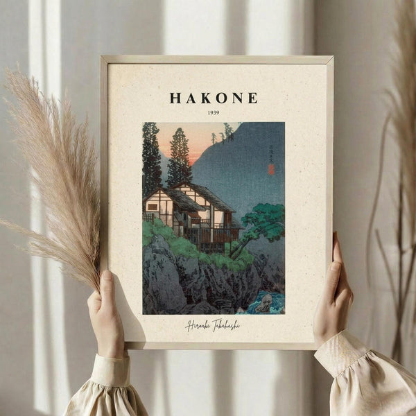 May include: Person in beige clothing holding framed vintage Japanese woodblock print showing traditional houses nestled among trees and rocky cliffs at twilight, with mountain silhouette in background. Light wooden frame against cream textured mat. Dried pampas grass visible in left hand. Soft natural lighting creates gentle shadows on neutral walls.