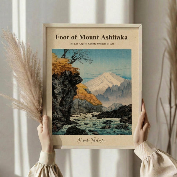 May include: Person in cream ruffled sleeves holding framed vintage Japanese woodblock print showing Mount Fuji landscape with flowing river, autumn foliage, and rocky terrain in muted blues, oranges and greens. Light wood frame against white curtained background with dried pampas grass visible to the left.