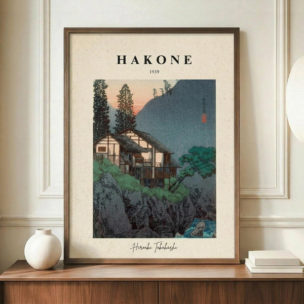 May include: Framed vintage Japanese woodblock print featuring traditional houses nestled among trees and rocky cliffs at twilight, with mountain silhouettes in background. Warm peachy-pink sky contrasts with deep blue hills. Print displays "HAKONE 1939" at top and artist signature below. Wooden frame sits on walnut credenza beside white ceramic vase and stacked books against cream walls with white trim molding.