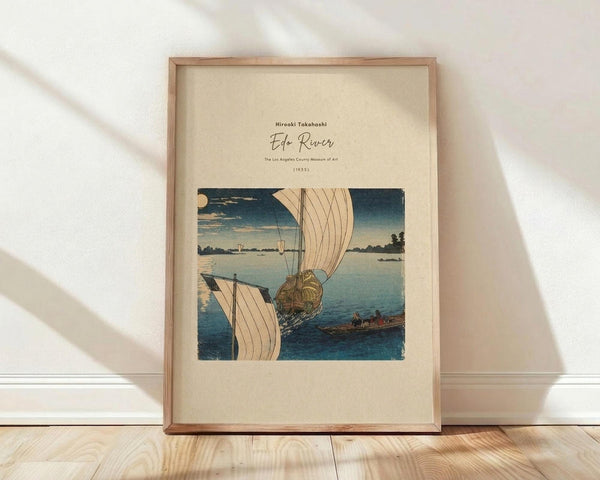 May include: Framed vintage Japanese woodblock print showing boats with white sails on blue water under evening sky with moon. Artwork features traditional ukiyo-e style with muted blues and beige tones. Light wooden frame with cream matting, leaning against white wall on natural wood floor with soft window light casting shadows.