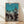 May include: Framed vintage Japanese woodblock print poster featuring silhouetted dancers in traditional clothing performing around a lantern-decorated festival structure. The artwork has a turquoise blue sky background fading to gray, with black silhouetted figures in dynamic dancing poses. The wooden frame sits on light wood flooring against a white wall with natural sunlight casting geometric shadows across the scene.