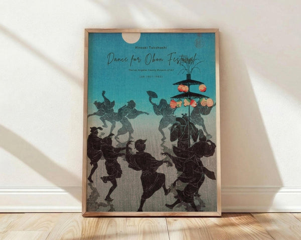 May include: Framed vintage Japanese woodblock print poster featuring silhouetted dancers in traditional clothing performing around a lantern-decorated festival structure. The artwork has a turquoise blue sky background fading to gray, with black silhouetted figures in dynamic dancing poses. The wooden frame sits on light wood flooring against a white wall with natural sunlight casting geometric shadows across the scene.