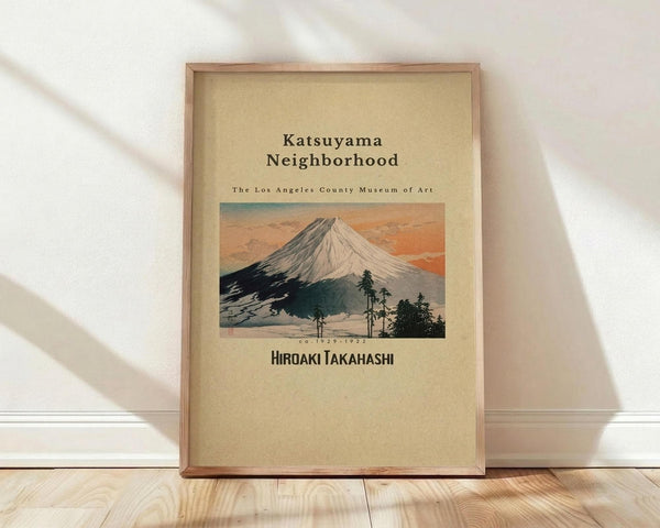 May include: A framed vintage Japanese woodblock print featuring Mount Fuji with snow-capped peak against an orange sunset sky, dark silhouetted trees in foreground. The artwork has cream-colored background with black text reading "Katsuyama Neighborhood" and "The Los Angeles County Museum of Art" above the image, and "Hiroaki Takahashi" below. Natural wood frame sits on light wooden floor against white wall with soft shadows.