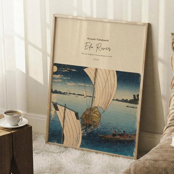 May include: Framed Japanese woodblock print poster showing boats with white sails on blue water under a twilight sky, displayed in light wooden frame leaning against white wall corner next to textured throw blanket and wooden side table with white coffee cup, creating cozy bedroom or living room setting with natural window lighting.