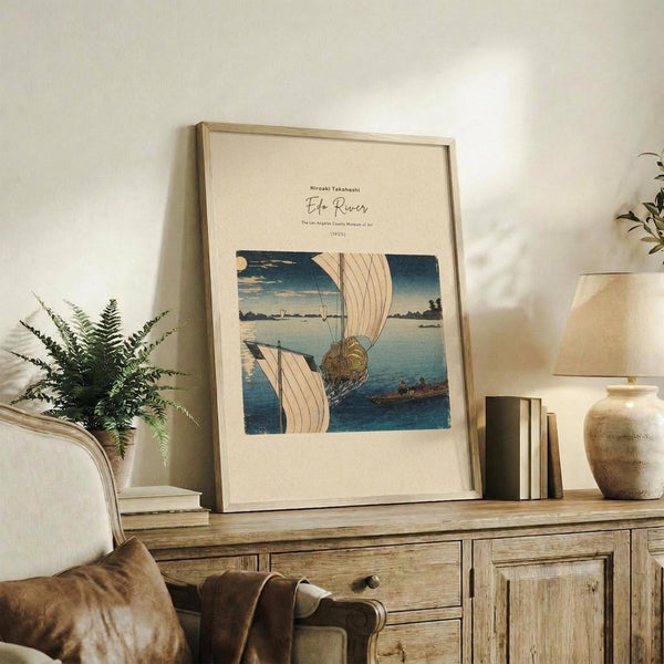 May include: Framed Japanese woodblock print showing boats with white sails on blue water, displayed on rustic wooden console table alongside fern plant, books, and ceramic pottery in neutral-toned living room with beige chair and soft natural lighting