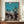 May include: Framed vintage Japanese festival poster showing silhouetted dancers in traditional clothing carrying colorful paper lanterns against a turquoise and cream gradient sky with full moon. The artwork displays on a wooden sideboard beside a white ceramic vase and stacked books in a room with white wainscoting and warm lighting.