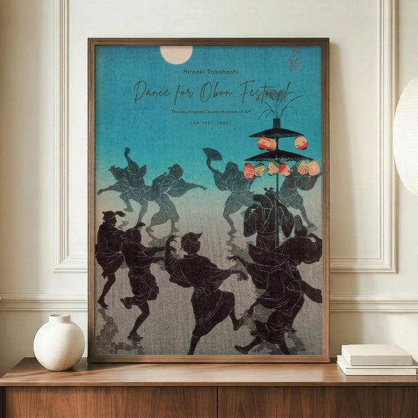 May include: Framed vintage Japanese festival poster showing silhouetted dancers in traditional clothing carrying colorful paper lanterns against a turquoise and cream gradient sky with full moon. The artwork displays on a wooden sideboard beside a white ceramic vase and stacked books in a room with white wainscoting and warm lighting.