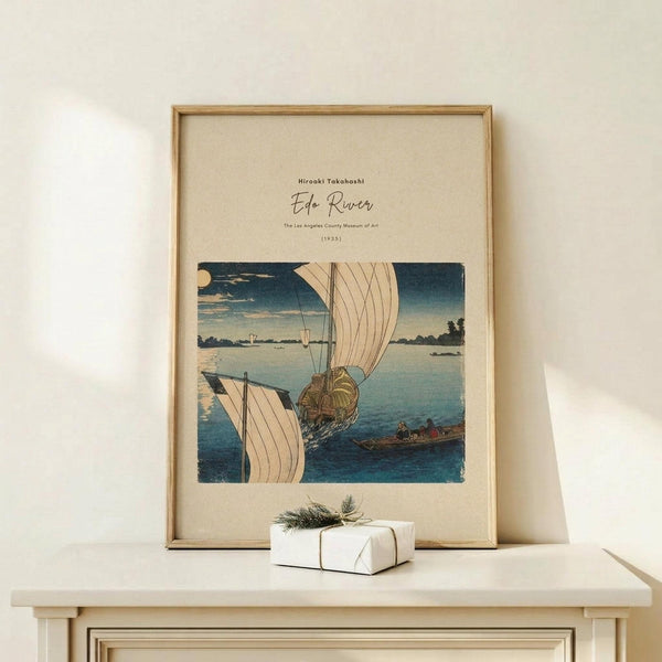 May include: Framed vintage Japanese woodblock print showing boats with white sails on blue water under evening sky with moon, displayed on light wood console table next to small wrapped gift with twine and greenery sprig against cream wall