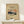 May include: Framed vintage-style Japanese art poster featuring Mount Fuji with snow-capped peak against orange sunset sky, displayed on light wood console table next to small white wrapped gift with greenery. Warm beige wall background, natural lighting, minimalist home decor styling.