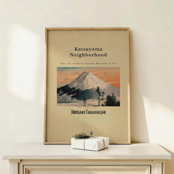 May include: Framed vintage-style Japanese art poster featuring Mount Fuji with snow-capped peak against orange sunset sky, displayed on light wood console table next to small white wrapped gift with greenery. Warm beige wall background, natural lighting, minimalist home decor styling.
