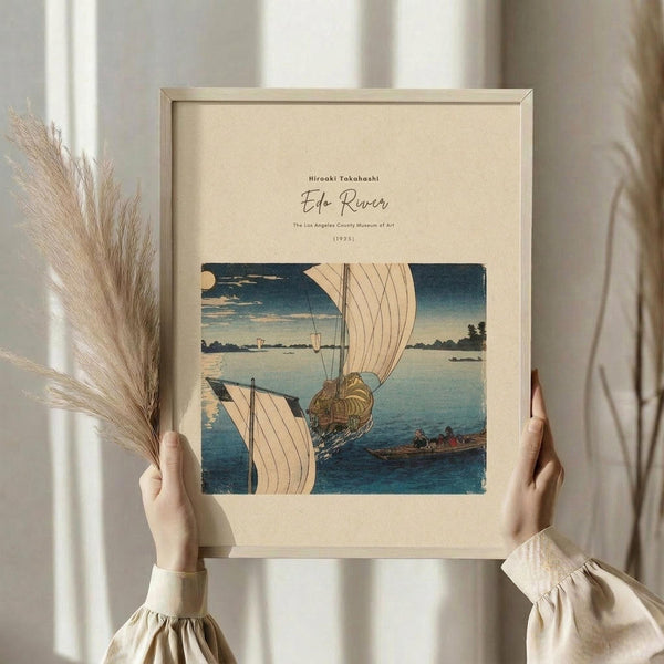 May include: Person holding framed vintage Japanese woodblock print showing sailing boats on blue river water with white sails, surrounded by calm landscape. Print features traditional ukiyo-e art style in blues, whites, and earth tones within light wood frame. Dried pampas grass visible beside frame against soft neutral curtained background.