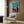 May include: Framed vintage Japanese festival poster with turquoise sky and silhouetted dancers carrying lanterns, displayed on wood-paneled wall above cream upholstered armchair with geometric pillow. Modern brass floor lamp and dark round side table with open book complete the cozy reading corner scene.
