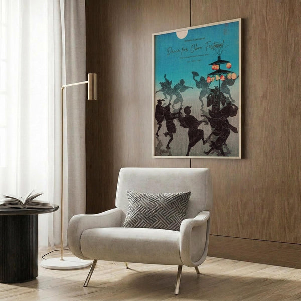 May include: Framed vintage Japanese festival poster with turquoise sky and silhouetted dancers carrying lanterns, displayed on wood-paneled wall above cream upholstered armchair with geometric pillow. Modern brass floor lamp and dark round side table with open book complete the cozy reading corner scene.