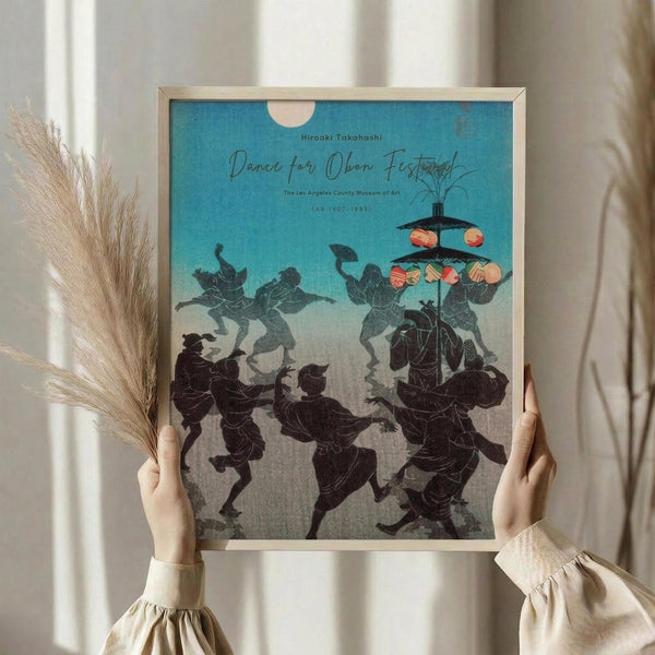 May include: Hands holding a framed vintage Japanese woodblock print showing silhouetted figures dancing around a tall lantern pole against a turquoise and cream gradient sky. The black figures appear to be celebrating, with colorful lanterns hanging from the pole. Dried pampas grass visible on the left side. Light wooden frame against soft neutral curtains and window light.