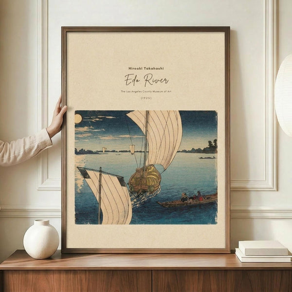 May include: Japanese woodblock print reproduction showing boats on blue water with white sails, displayed in brown wooden frame on wooden sideboard. Person's hand touches frame edge. White ceramic vase and stacked books nearby. Cream colored walls with white molding in background. Print features traditional ukiyo-e style with blue tones and beige matting.