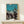 May include: Framed vintage Japanese woodblock print showing silhouetted figures in traditional dress dancing and celebrating, with lanterns and turquoise sky gradient background. Displayed on light wood console table beside wrapped gift with evergreen sprig. Warm neutral wall backdrop with natural lighting.