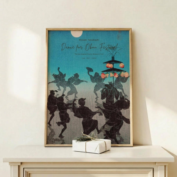 May include: Framed vintage Japanese woodblock print showing silhouetted figures in traditional dress dancing and celebrating, with lanterns and turquoise sky gradient background. Displayed on light wood console table beside wrapped gift with evergreen sprig. Warm neutral wall backdrop with natural lighting.