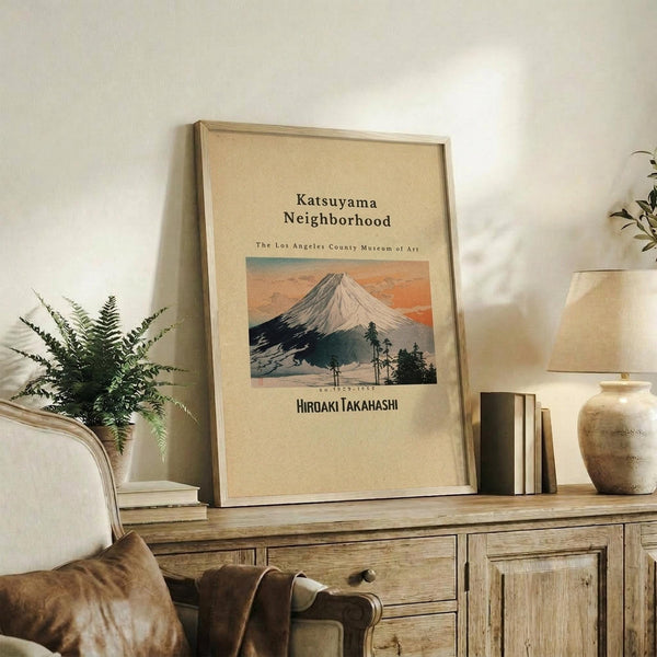 May include: Framed vintage Japanese art print featuring Mount Fuji with snow-capped peak against orange sunset sky, displayed on rustic wooden console table alongside fern plant, books, and ceramic vase in neutral-toned living room setting with cream upholstered chair and white walls