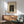May include: Japanese woodblock print poster in wooden frame hanging above mid-century modern credenza with ceramic lamp. Green upholstered armchair with wood frame sits nearby. Print shows figure in dark robes with colorful pottery vessels. Warm sunlight creates shadows across white wall. Room has minimalist aesthetic with natural wood tones.