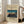 May include: Framed vintage Japanese woodblock print showing traditional sailing junks anchored in a bay at twilight, with blue-orange gradient sky and crescent moon. Print features beige museum-style matting and thin wooden frame, leaning against white wall next to beige upholstered chair in bright, naturally-lit room with hardwood floors.