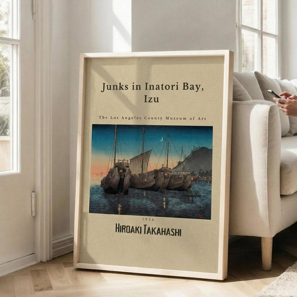 May include: Framed vintage Japanese woodblock print showing traditional sailing junks anchored in a bay at twilight, with blue-orange gradient sky and crescent moon. Print features beige museum-style matting and thin wooden frame, leaning against white wall next to beige upholstered chair in bright, naturally-lit room with hardwood floors.