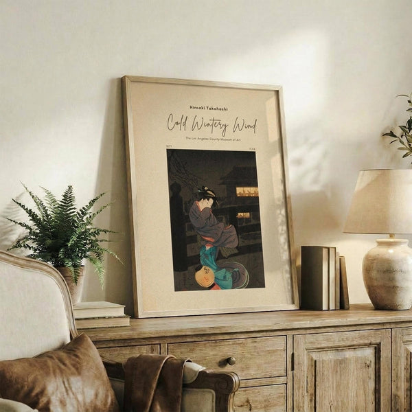 May include: Framed Japanese woodblock print poster displayed on rustic wooden credenza alongside fern plant, stacked books, and ceramic vase. Print shows woman in traditional dress kneeling by warming brazier in muted earth tones. Natural lighting creates warm shadows across cream-colored wall and neutral living room setting.