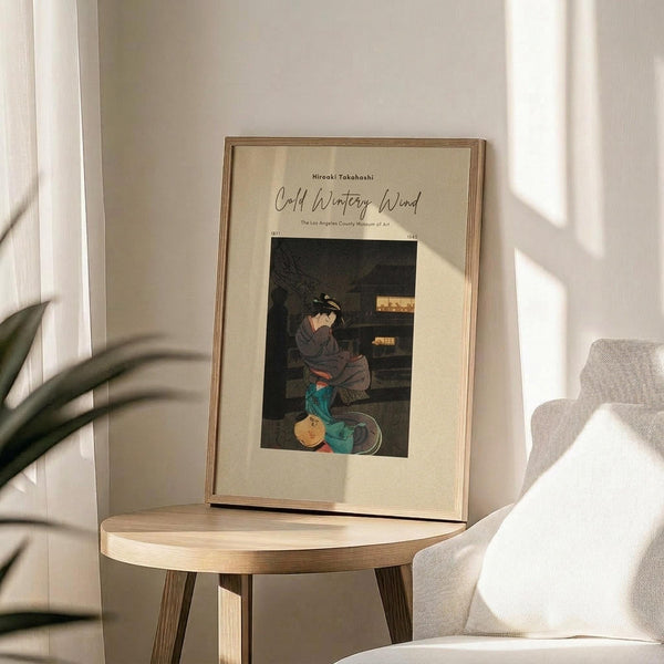 May include: Framed vintage Japanese woodblock print poster displayed on round wooden side table next to beige upholstered chair. Artwork shows figure in dark robes kneeling beside turquoise ceramic vessels. Cream matting in thin wooden frame. Soft natural lighting creates window shadows on white wall. Green plant leaves visible in corner.