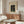 May include: Japanese woodblock print poster in beige mat and thin frame, hanging above modern walnut credenza in minimalist living room. Print shows woman in traditional dress sitting by wooden structure. White sculptural chair, glass oval coffee table, and spherical table lamp visible. Warm wood tones throughout neutral-colored space with natural lighting.