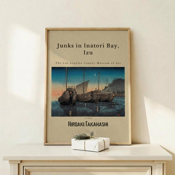 May include: Framed vintage Japanese woodblock print showing traditional sailing junks on blue water at twilight, displayed on light wood table with small wrapped gift. Print features boats with tall masts against orange sunset sky and mountains, presented in natural wood frame on neutral wall background.
