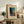 May include: Framed vintage Japanese woodblock print showing a figure carrying bundles at dusk, with dark silhouetted trees against a turquoise sky. The beige matted artwork sits on a rustic wooden sideboard alongside a fern plant, ceramic vase, and books in a warmly lit living room with cream upholstered furniture.
