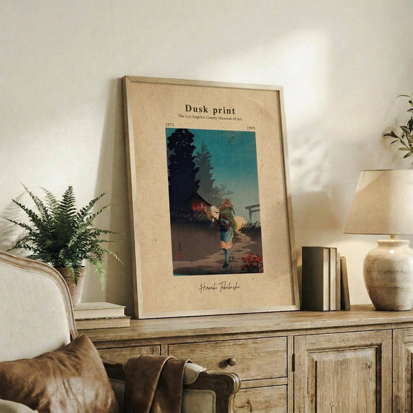 May include: Framed vintage Japanese woodblock print showing a figure carrying bundles at dusk, with dark silhouetted trees against a turquoise sky. The beige matted artwork sits on a rustic wooden sideboard alongside a fern plant, ceramic vase, and books in a warmly lit living room with cream upholstered furniture.