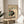 May include: Framed vintage-style Japanese artwork showing a black and white cat sitting beneath a tomato plant with red tomatoes and green leaves, displayed on a round wooden side table next to a beige upholstered chair with a cream throw pillow, natural lighting creating soft shadows on white walls with a plant visible in the corner