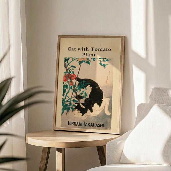 May include: Framed vintage-style Japanese artwork showing a black and white cat sitting beneath a tomato plant with red tomatoes and green leaves, displayed on a round wooden side table next to a beige upholstered chair with a cream throw pillow, natural lighting creating soft shadows on white walls with a plant visible in the corner