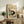 May include: Framed vintage Japanese-style artwork showing a black and white cat sitting beside a tomato plant with red tomatoes and green leaves, displayed on a rustic wooden sideboard alongside fern plant, ceramic vase, and books in a neutral-toned living room with beige chair visible.