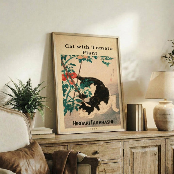 May include: Framed vintage Japanese-style artwork showing a black and white cat sitting beside a tomato plant with red tomatoes and green leaves, displayed on a rustic wooden sideboard alongside fern plant, ceramic vase, and books in a neutral-toned living room with beige chair visible.