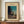 May include: Framed vintage Japanese woodblock print showing a figure carrying bundles walking on a path at dusk, with traditional buildings and trees silhouetted against a turquoise to peach gradient sky. The cream-colored matted print displays in a brown wooden frame on a walnut sideboard beside a white ceramic vase and stacked books against white wainscoting.