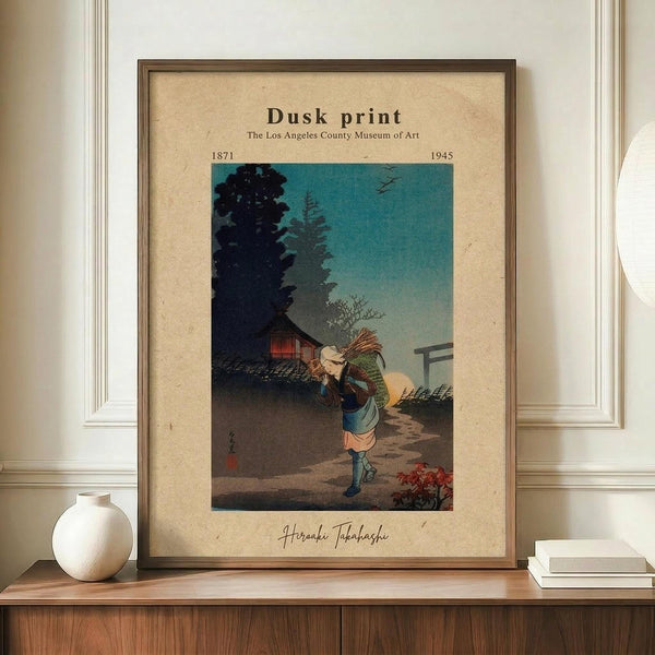 May include: Framed vintage Japanese woodblock print showing a figure carrying bundles walking on a path at dusk, with traditional buildings and trees silhouetted against a turquoise to peach gradient sky. The cream-colored matted print displays in a brown wooden frame on a walnut sideboard beside a white ceramic vase and stacked books against white wainscoting.