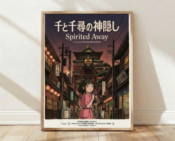 May include: Framed Spirited Away movie poster featuring Chihiro in pink outfit standing on traditional Japanese street with multi-story bathhouse building in background. Poster has Japanese text at top with English title below. Warm sunset lighting with paper lanterns and traditional architecture. Light wooden frame displayed against white wall on wooden floor with soft shadows.