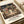 May include: Spirited Away movie poster print lying flat on white paper with cream ribbon nearby. Shows anime girl with short black hair in white and green striped shirt standing on wooden steps of traditional Japanese bathhouse at night. Detailed illustration features multi-story building with red and green architectural details, glowing lanterns, and Japanese text. Rich colors include deep blues, warm browns, and golden lighting.