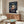 May include: Framed Spirited Away movie poster mounted on wall above wooden sideboard in modern living room. Poster features anime girl character Chihiro in red outfit with train and floating spirits on dark background. White upholstered chair, glass coffee table, and table lamp visible in minimalist interior setting with neutral beige walls.