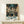 May include: Framed Spirited Away movie poster displayed on light wood shelf against white wall. Features young girl character Chihiro in traditional clothing walking through bustling Japanese street scene with lanterns, food stalls, and other characters. Artwork shows Studio Ghibli's distinctive animation style with rich greens, blues, and warm lighting. Small white decorative box sits beside the wooden-framed print on the minimalist shelf.