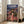 May include: Framed Spirited Away movie poster leaning against white wall in bright living room. Features young girl in red outfit on wooden bridge with ornate multi-story bathhouse building behind her at sunset. Japanese text at top with English title below. Warm lighting from large windows, beige sofa visible on right, light wood floors.
