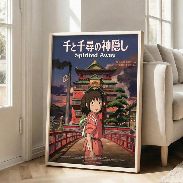 May include: Framed Spirited Away movie poster leaning against white wall in bright living room. Features young girl in red outfit on wooden bridge with ornate multi-story bathhouse building behind her at sunset. Japanese text at top with English title below. Warm lighting from large windows, beige sofa visible on right, light wood floors.