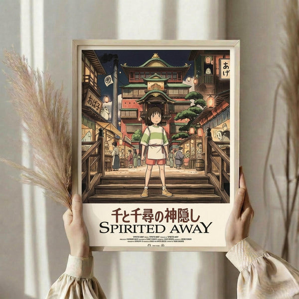 May include: Hands holding a framed Spirited Away movie poster showing the young protagonist Chihiro standing in a traditional Japanese bathhouse street scene at night. The artwork features detailed anime-style illustration with warm lantern lighting, traditional architecture, and spirit characters. Light wood frame against white curtained background with dried pampas grass visible to the left.