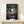 May include: Framed Spirited Away movie poster featuring Chihiro in red top with small white spirits floating around her, train tracks below, displayed on light wood shelf beside small wrapped gift with dried sprig on white wall