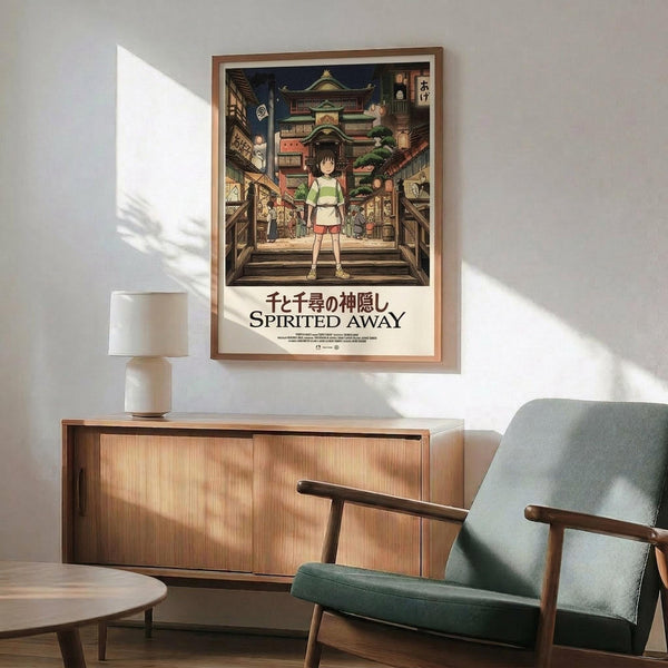 May include: Framed Spirited Away movie poster displayed on white wall above wooden credenza with white ceramic lamp. Green upholstered mid-century modern chair with wooden arms sits nearby. Poster shows young girl in green shirt standing in traditional Japanese bathhouse street scene with colorful buildings, warm lighting, and Japanese text. Natural sunlight creates diagonal shadows across the minimalist room setting.