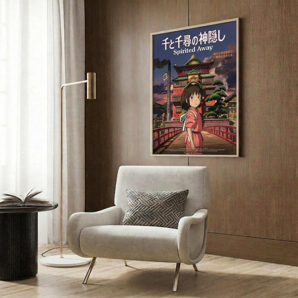 May include: Spirited Away anime poster with Japanese text and English title, featuring a young girl in pink kimono standing before a traditional multi-story bathhouse building under dramatic sky. Poster shown framed on wood-paneled wall above modern beige armchair with geometric patterned pillow. Contemporary room setting with gold floor lamp and side table with open book.