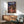 May include: Framed Studio Ghibli Spirited Away movie poster hanging on white wall above wooden sideboard with ceramic lamp. Poster shows anime girl in red outfit standing on wooden bridge before ornate Japanese bathhouse building against twilight sky. Mid-century modern room setting with teal upholstered armchair, round wooden coffee table, and natural sunlight casting geometric shadows across the scene.