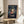 May include: Framed Spirited Away movie poster featuring anime girl character Chihiro in red kimono against dark blue background with train and floating spirits, displayed on round wooden side table next to beige upholstered chair and plant in bright sunlit room with white walls